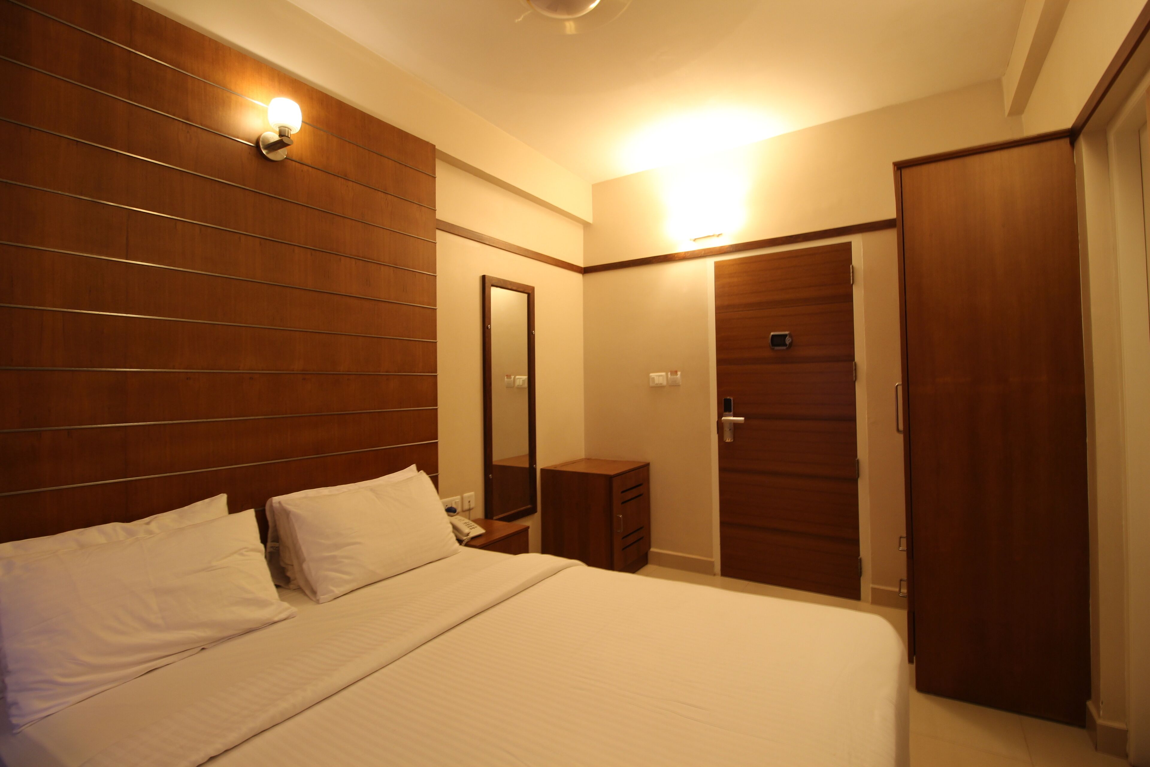 A hotel room with a large bed, wooden headboard, a bedside table with a lamp, a mirror, and a wardrobe.