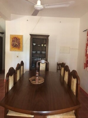 A dining room with a large wooden table, six chairs, a ceiling fan, a decorative painting, and a cabinet with dishes.