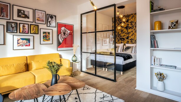 Una sala de estar moderna con un sofá amarillo, una mesa de centro de madera y un separador de vidrio que da a un dormitorio.
