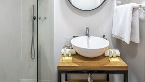 Un baño moderno con ducha envuelta en vidrio, un espejo redondo, tocador de madera y lavamanos blanco.