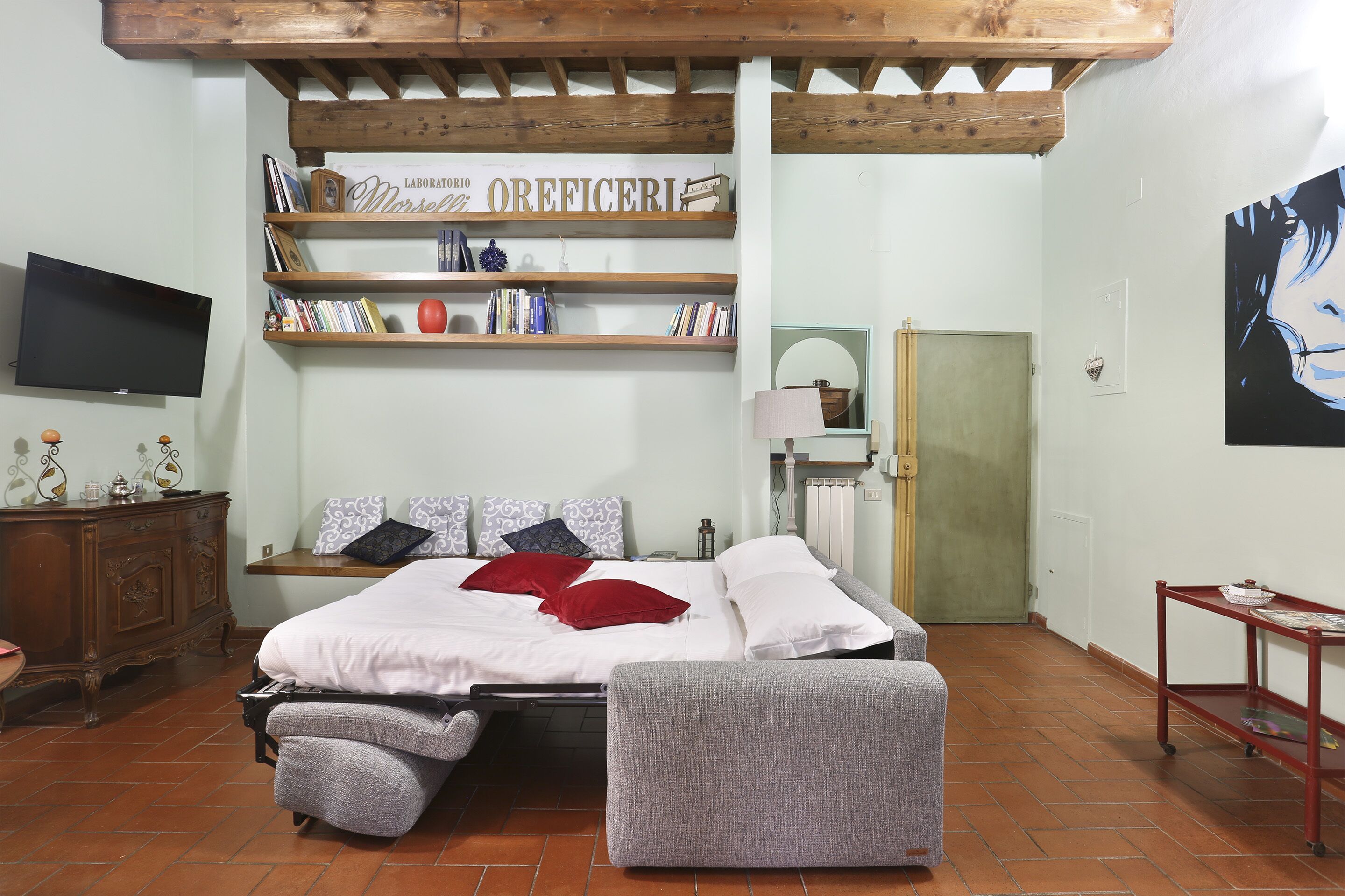 A bedroom with a bed, a wooden shelf with books, a TV, and a painting on the wall.