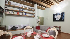 Zona pranzo con tavolo in legno apparecchiato per un pasto, libreria con libri e oggetti decorativi e un quadro appeso al muro.