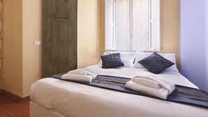 Un letto rifatto con lenzuola bianche e due cuscini scuri a motivi decorativi, una finestra con persiane chiuse e una porta con stipite in legno.