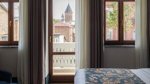 A hotel room with a large bed, a blue chair, and a view of a building with a tower through a window. - Boutique Hotel Puris Moedani (Tbilisi)