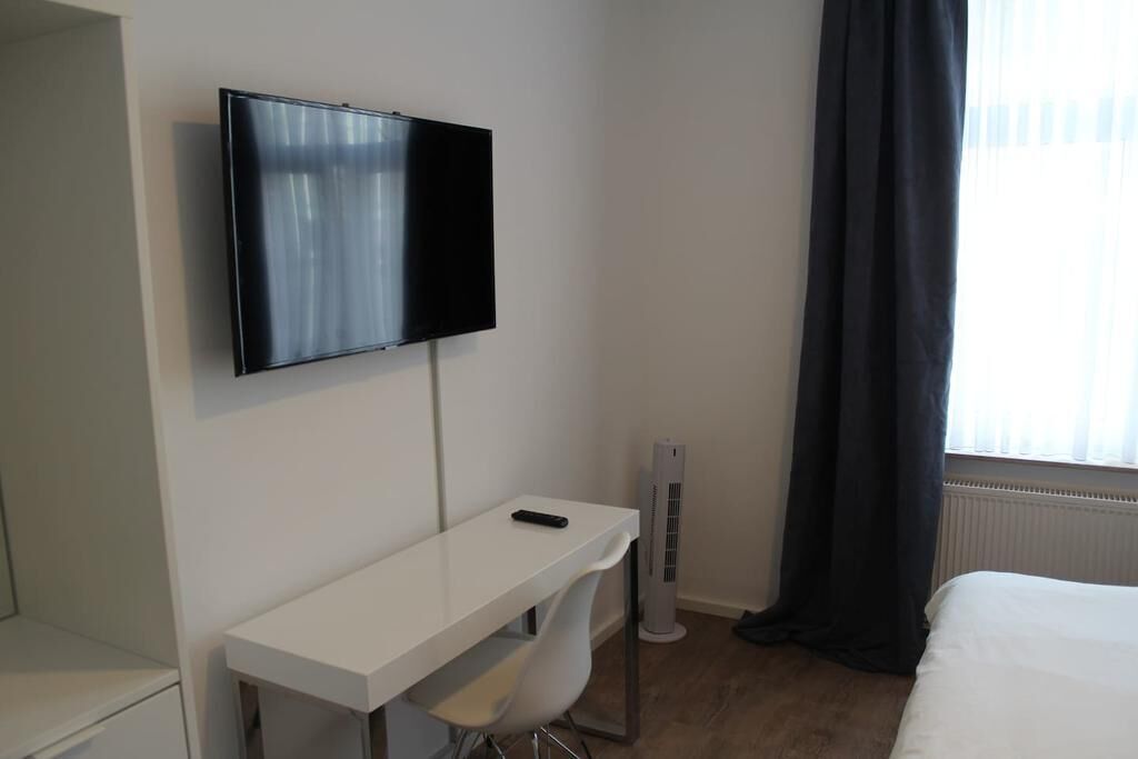 Une chambre d’hôtel équipée d’un téléviseur à écran plat fixé au mur, d’un bureau blanc avec une chaise, d’un radiateur et d’un lit recouvert d’une couette blanche.