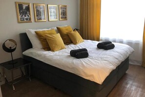 A neatly made bed with white and mustard pillows, two rolled towels, and a framed picture collection on the wall. - Chillinghouse 9 (Siegburg)