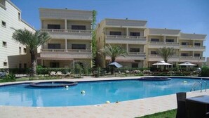 A swimming pool with a tiled deck, surrounded by lounge chairs and umbrellas, with a multi-story building in the background. - Muhanna Chalets (Al Khiran)
