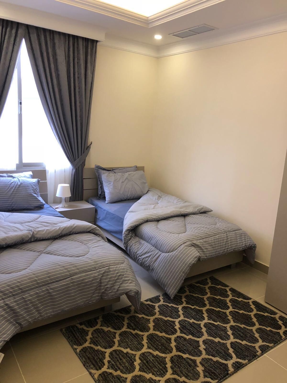 Un dormitorio con dos camas, una mesita de noche, una lámpara y una alfombra estampada.