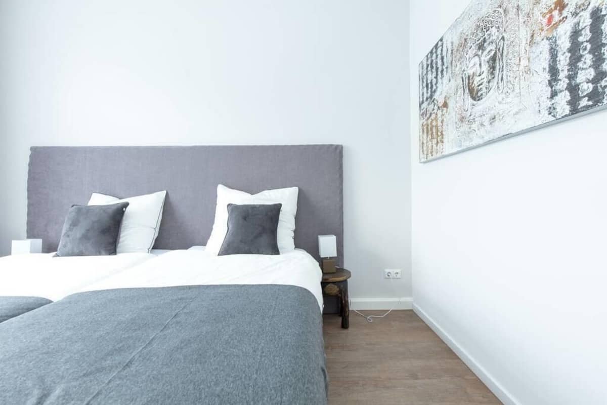 Une chambre à coucher moderne avec une tête de lit grise, deux oreillers gris, une couvre-lit blanche et une table de chevet en bois avec une lampe.