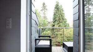 A balcony with two black cushioned seating areas and a small table, overlooking a lush green landscape with tall trees. - Chillinghouse 11 (Siegburg)