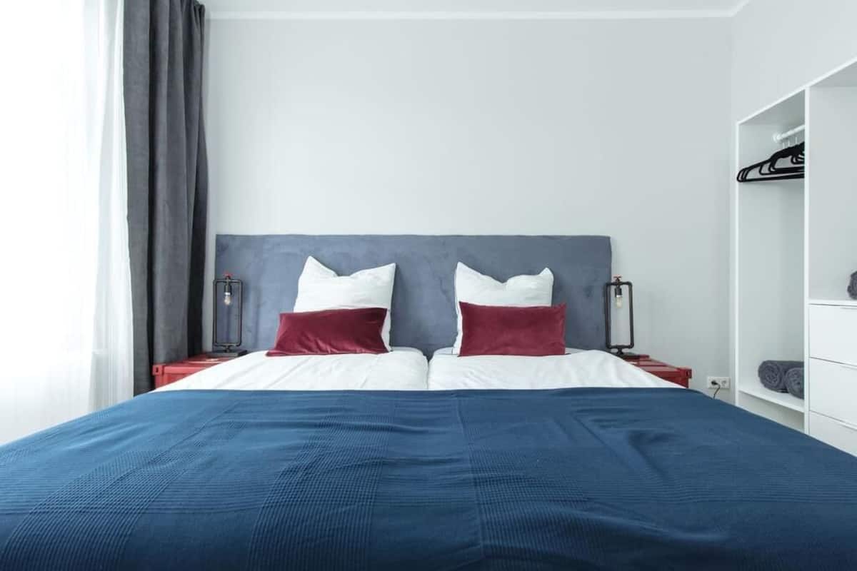Un lit bien fait, avec des oreillers blancs et rouges, une couverture bleue et une tête de lit grise. On y voit une table de chevet blanche avec une lampe et une valise rouge posée dessus. Un rideau gris est partiellement tiré sur le côté.