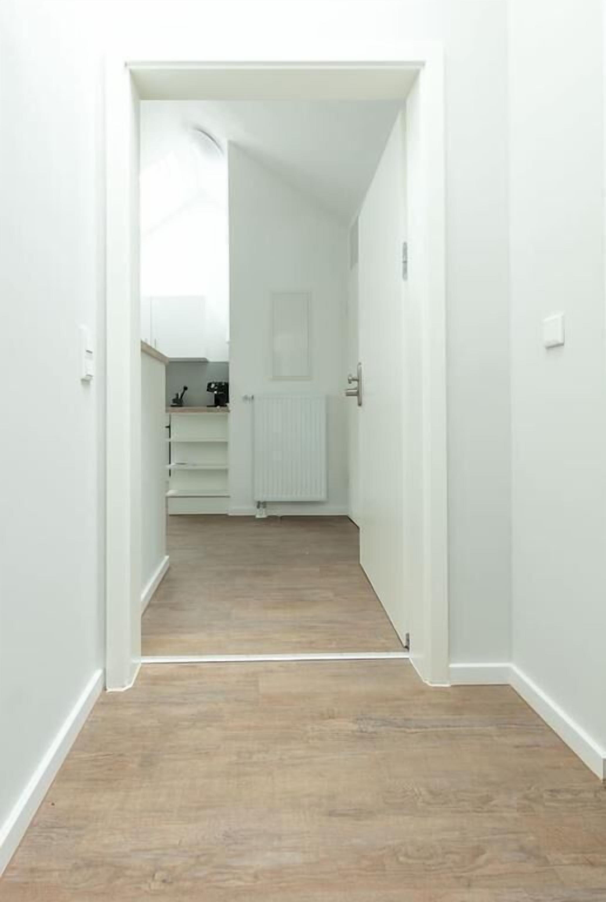 Un pasillo estrecho con paredes blancas y suelo de madera que conduce a una habitación con un radiador y una puerta.