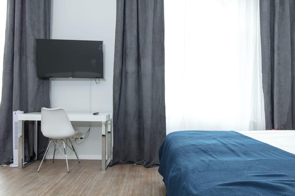 Hotellihuone, jossa on sänky, työpöytä tuolilla, seinälle asennettu taulutelevisio ja harmaat verhot.