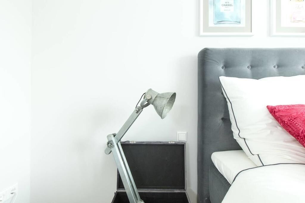 Ein ordentlich bezogenes Bett mit weißen und roten Kissen, einem grauen Kopfteil und einem grauen Nachttisch mit einer Lampe.