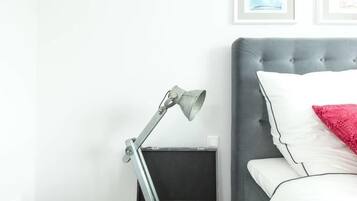 Ein ordentlich bezogenes Bett mit weißen und roten Kissen, einem grauen Kopfteil und einem grauen Nachttisch mit einer Lampe.