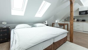 Quarto moderno com teto inclinado, claraboias, estrutura de cama de madeira, mesa de cabeceira com abajur e uma kitchenette ao fundo.