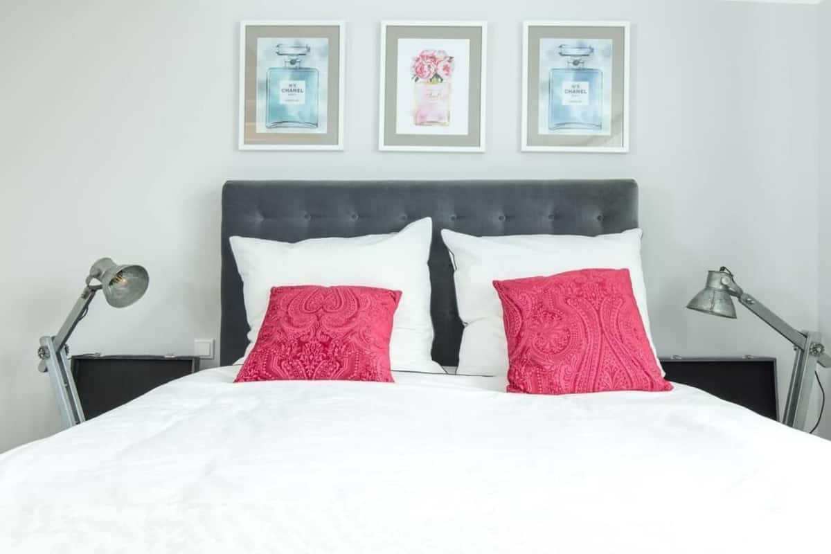 Un lit bien fait, avec des oreillers blancs et roses à motifs, une tête de lit sombre et trois tableaux encadrés accrochés au mur.