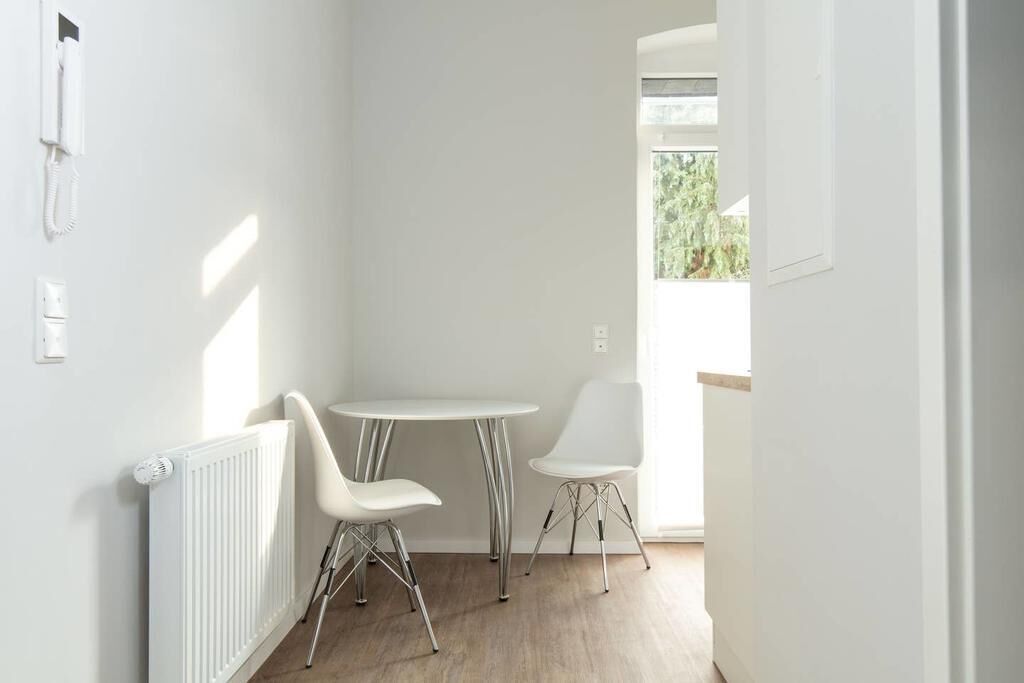 Een lichte, kleine kamer met een witte radiator, twee witte stoelen, een klein, rond tafeltje en een raam met uitzicht op groen.