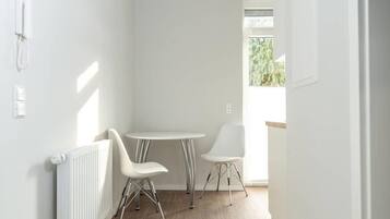 Een lichte, kleine kamer met een witte radiator, twee witte stoelen, een klein, rond tafeltje en een raam met uitzicht op groen.