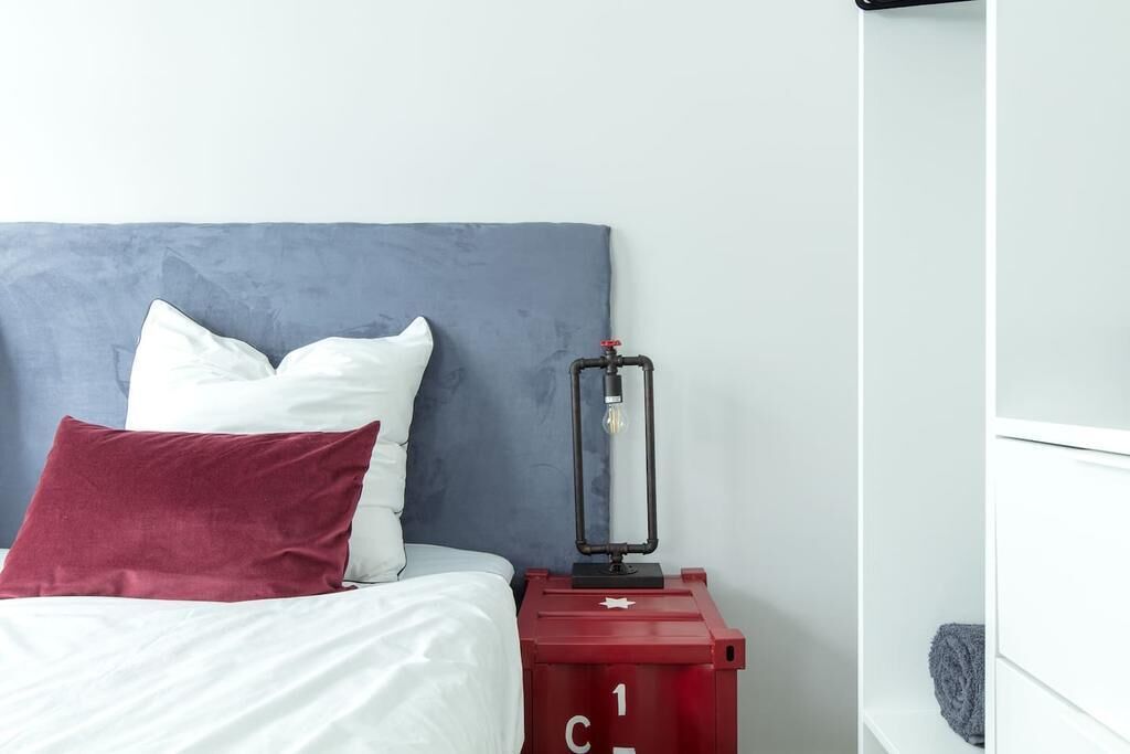 Ein Bett mit grauem Kopfteil, weißen und burgunderfarbenen Kissen sowie einem Nachttisch in Rot mit einer Lampe.