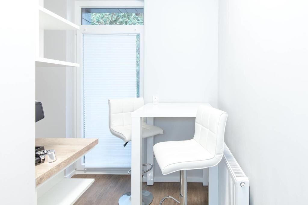 Una habitación pequeña y blanca con un escritorio, dos sillas, un radiador y una ventana con persianas.