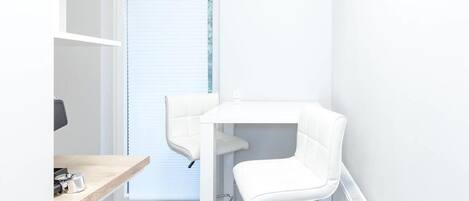 Una habitación pequeña y blanca con un escritorio, dos sillas, un radiador y una ventana con persianas.