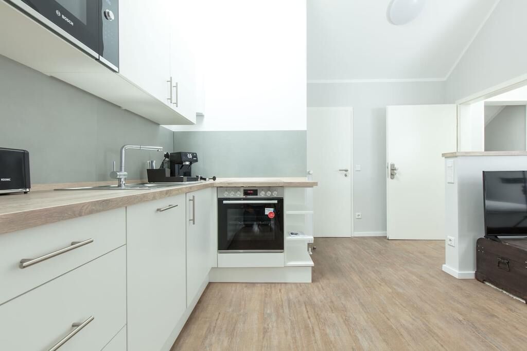 A compact kitchen with white cabinets, a built-in oven, a coffee maker, and a television on a wooden cabinet.