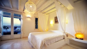 A bedroom with a large bed, a canopy, a bedside lamp, and a view of the sea through the window. - Villa Akoya III by The Pearls Collection (Mykonos)