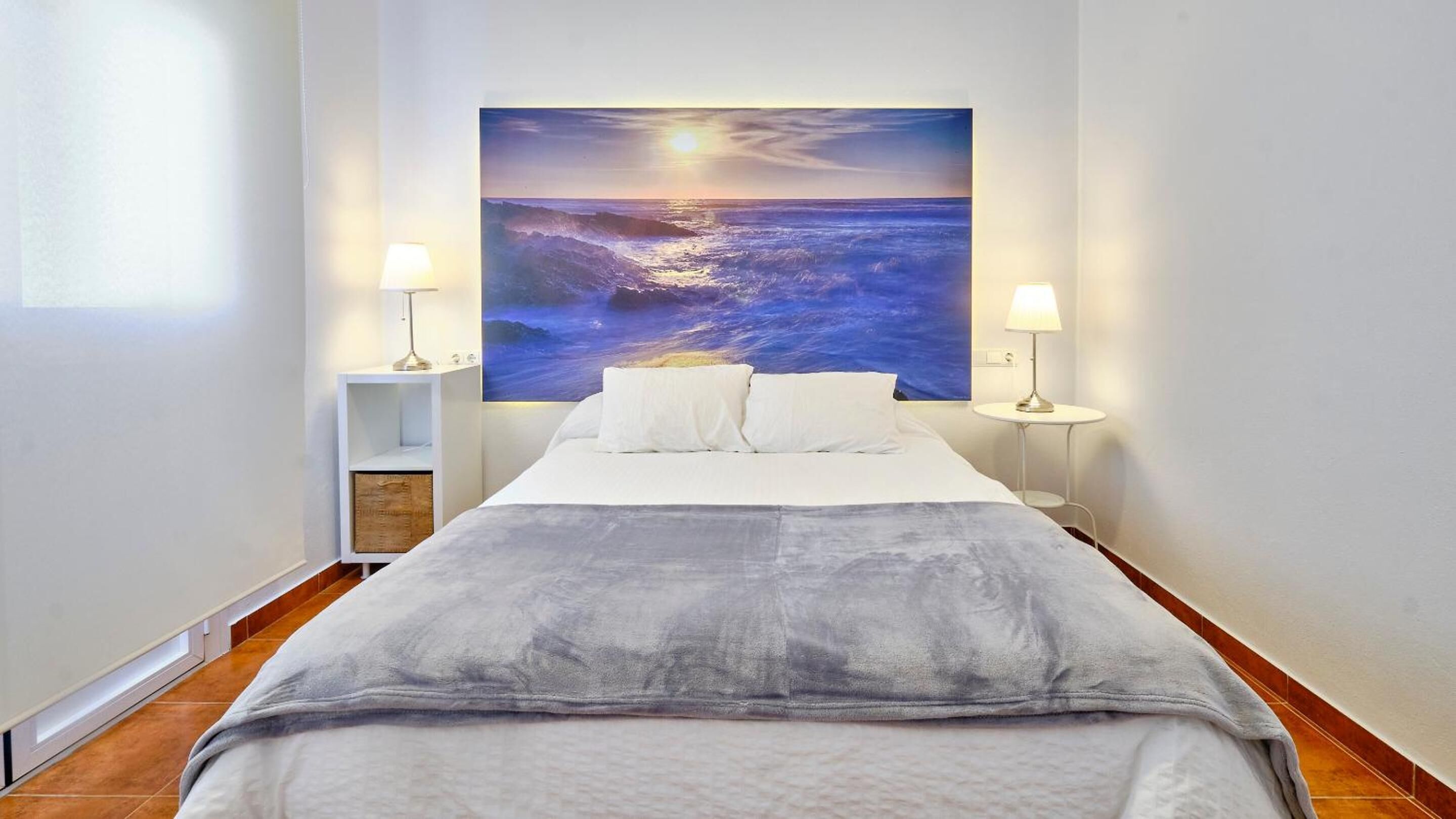 Un dormitorio con una pintura de una amplia vista al océano, una cama con ropa blanca y dos lámparas de noche.