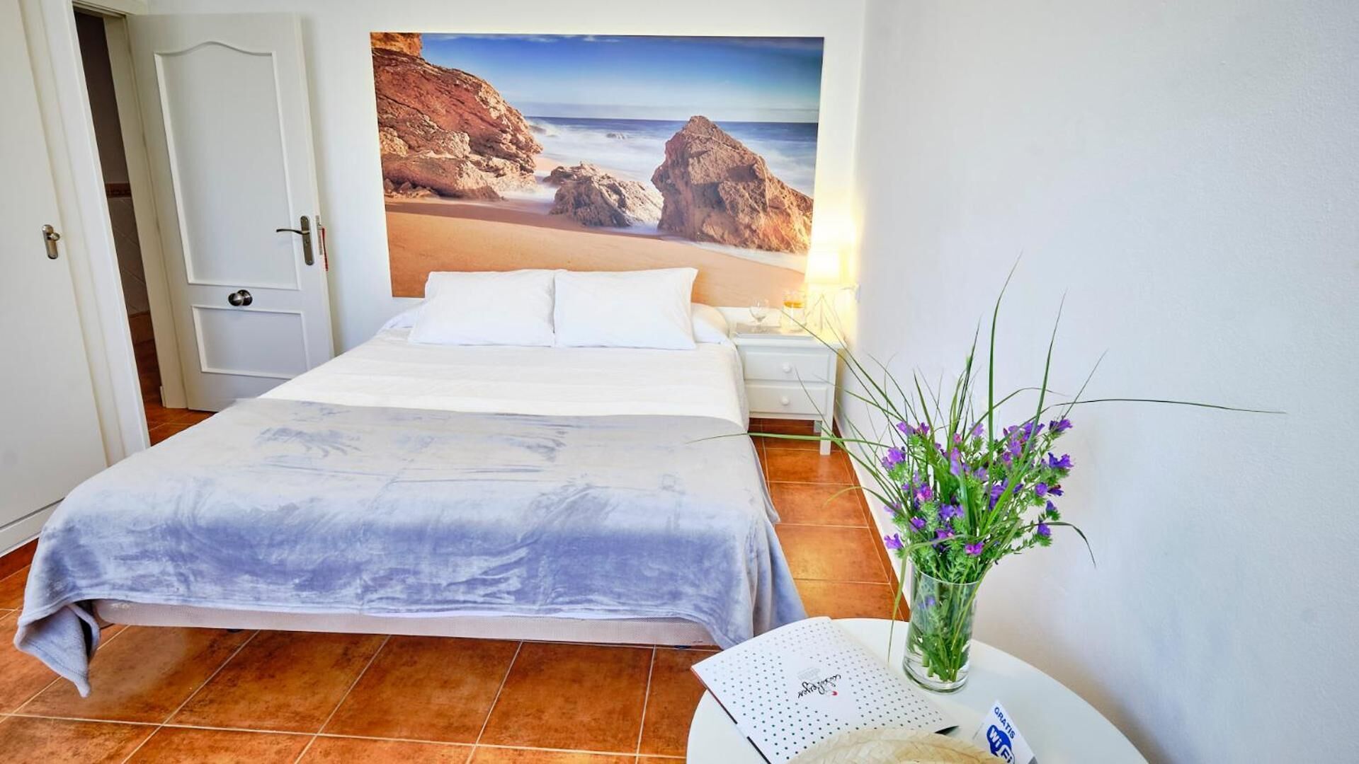 A bedroom with a bed, a nightstand, a vase with flowers, and a poster of a beach scene.