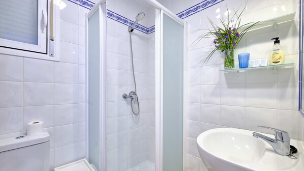 A bathroom with white tiles, a shower, a toilet, a sink, and a shelf with toiletries.