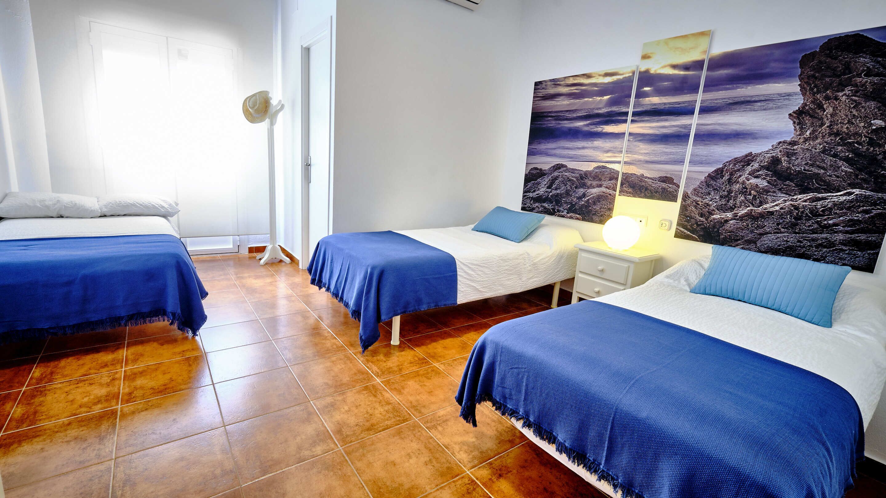 A room with two beds, a nightstand with a lamp, a large painting of a sunset, and a white door.