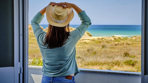 A person in a green top and jeans, holding a straw hat, looking out at a beach with sand dunes and the ocean.