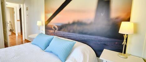 A hotel room with a large bed, two bedside tables, a nightstand, a wall-mounted lamp, and a large painting of a bridge.