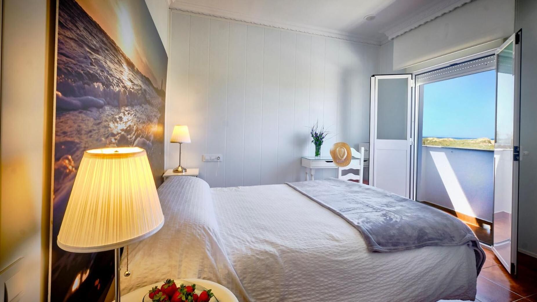 Habitación de hotel con cama, mesita de noche, lámpara, un florero con flores y vista al mar a través de una puerta corrediza.