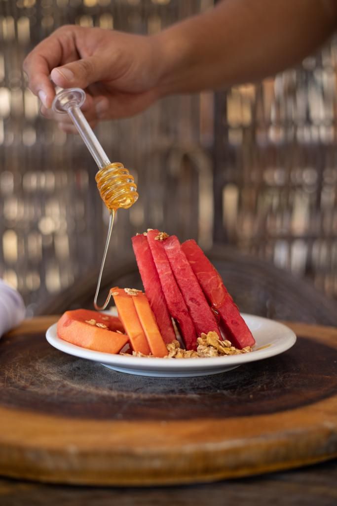 A plate with sliced fruits and granola, a honey dipper with honey, and a hand holding it.