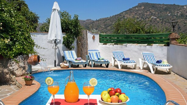 Ein Poolbereich mit Liegestühlen, einem Tisch mit Getränken und Früchten sowie Blick auf Hügel.