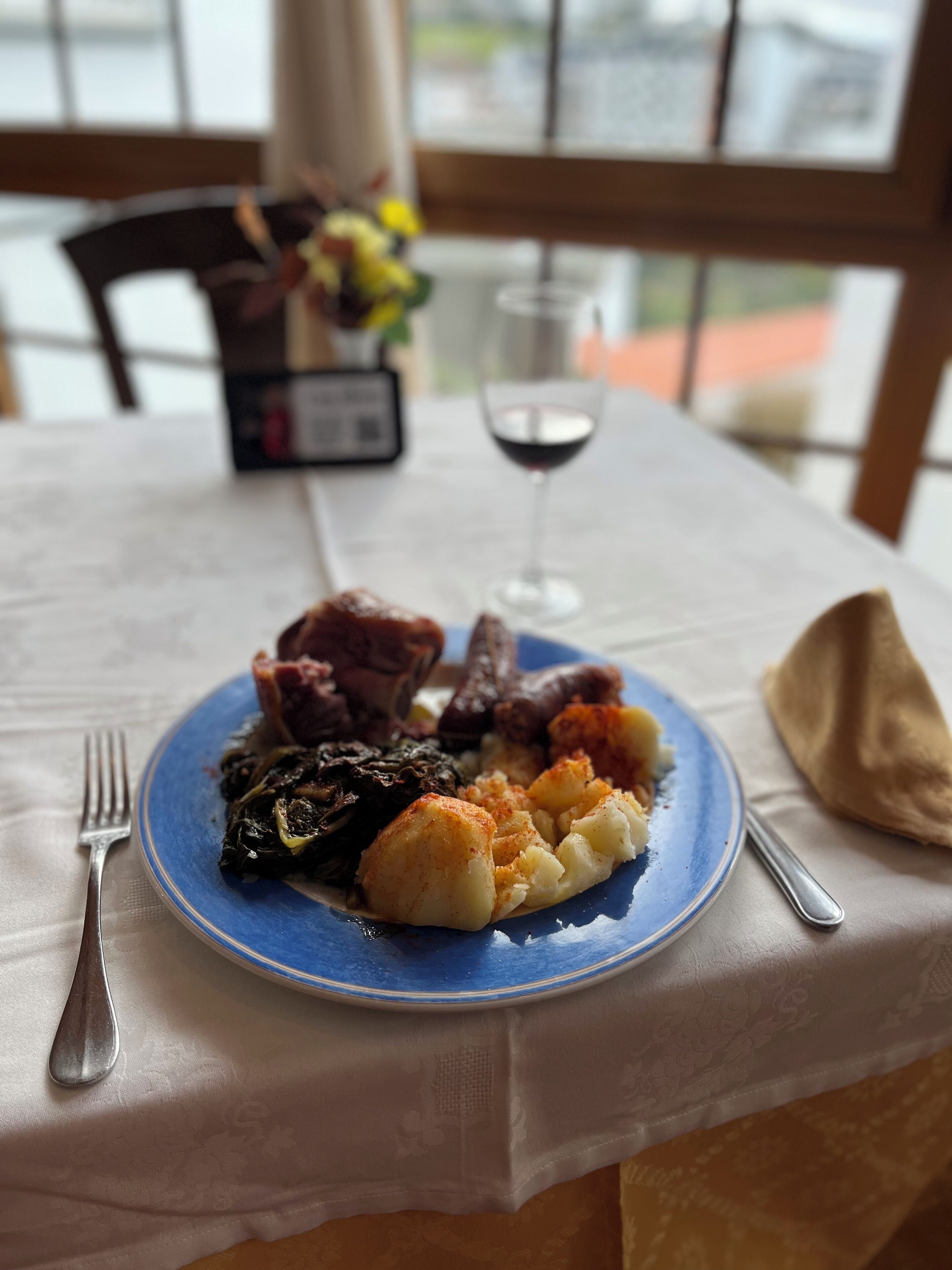 A plate of food on a table with a fork and spoon, a wine glass, and a chair in the background.