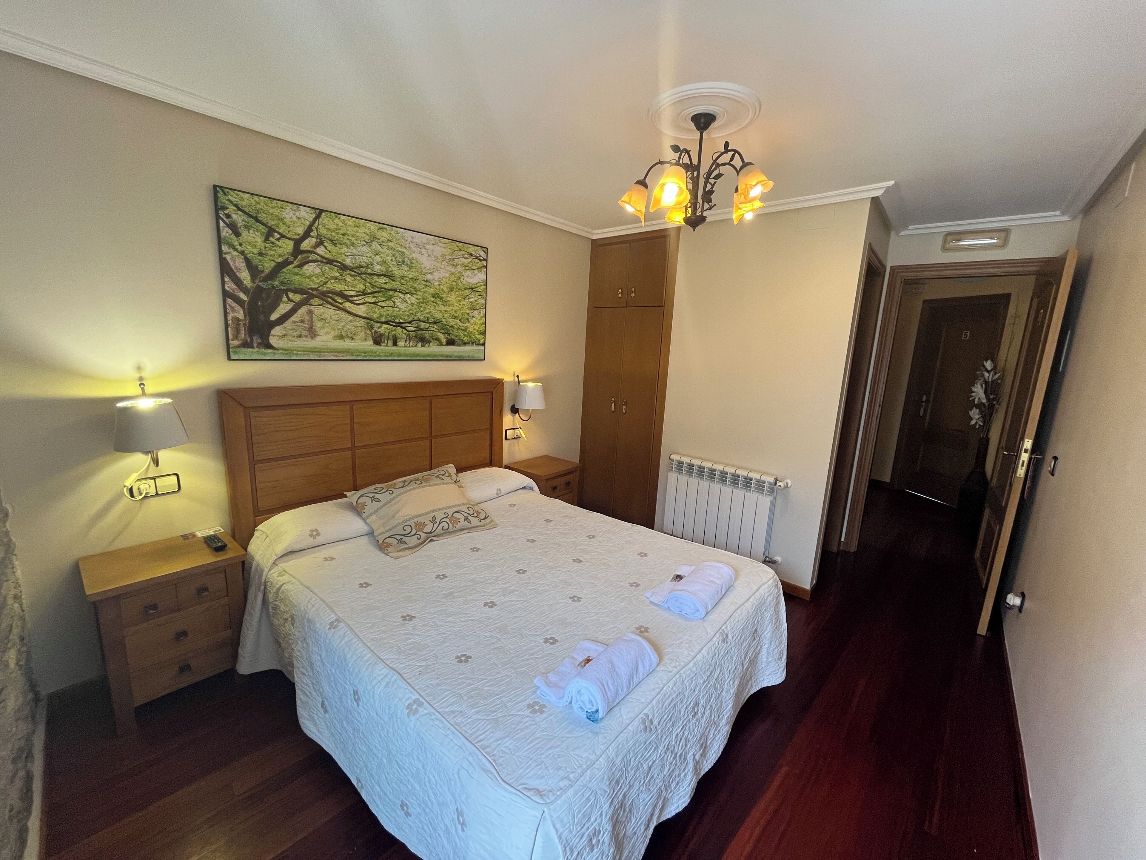 A neatly made bed with a floral bedspread, a nightstand with a lamp, a painting of a tree on the wall, and a wooden wardrobe.