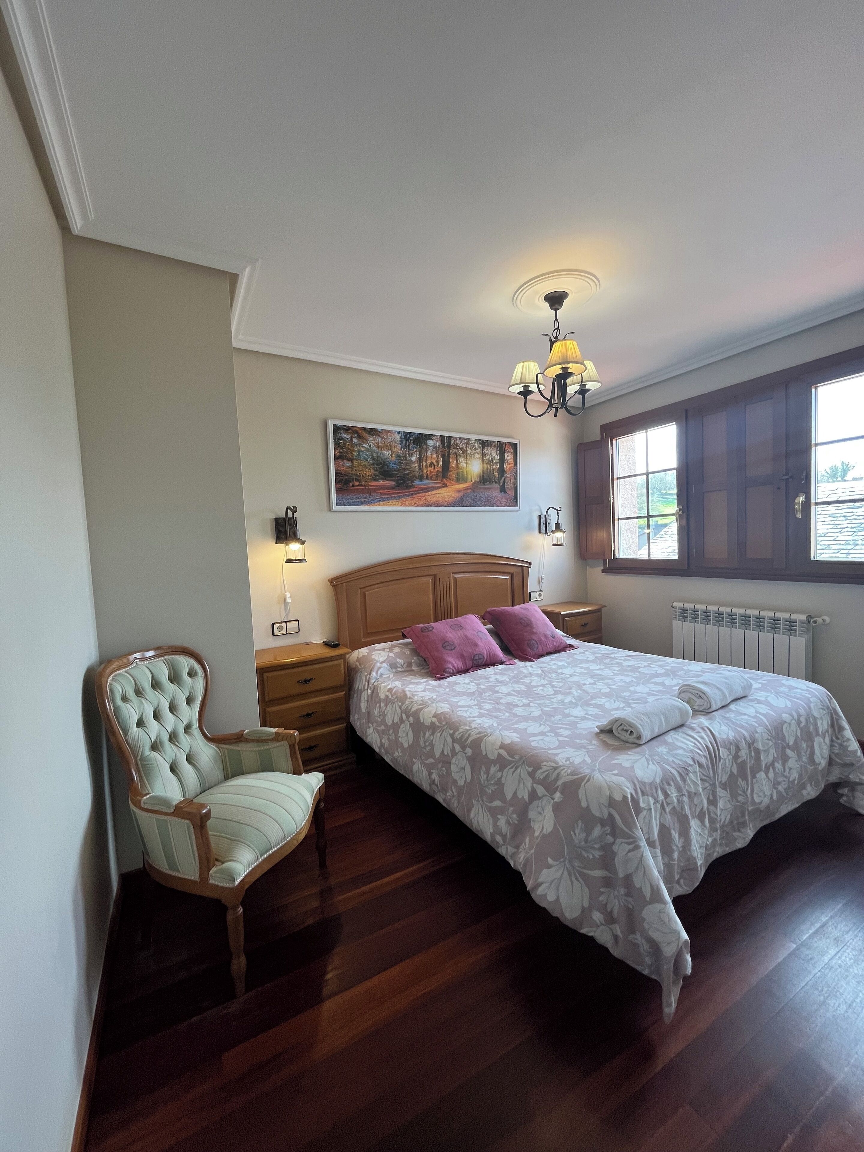 A bedroom with a bed, a chair, a nightstand, a lamp, a painting, and a window.