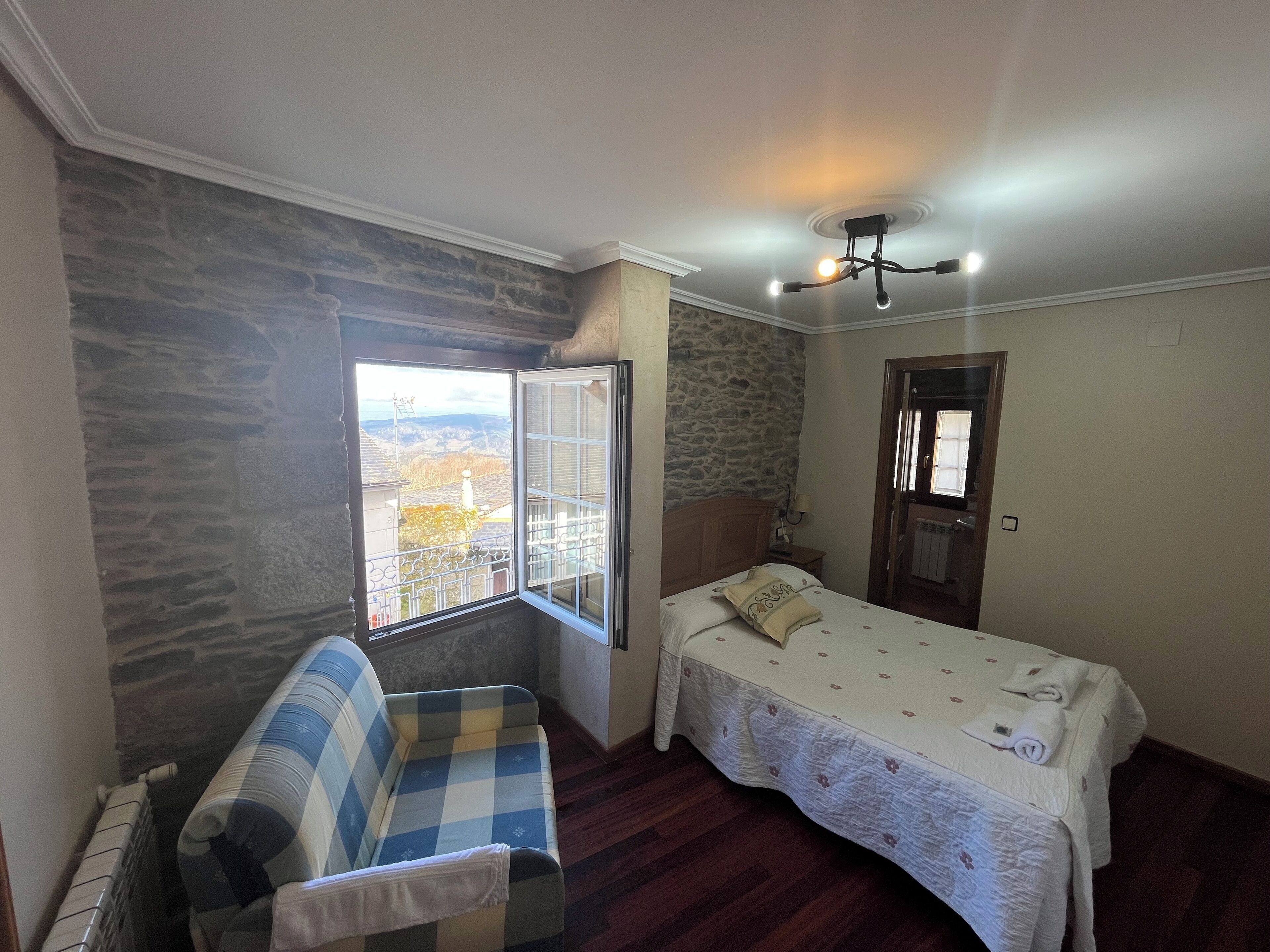 Ein Schlafzimmer mit Steinwand, einem Fenster mit Aussicht, einem Bett mit weißer Bettdecke, einem blau-weiß karierten Sofa und einem Deckenventilator.