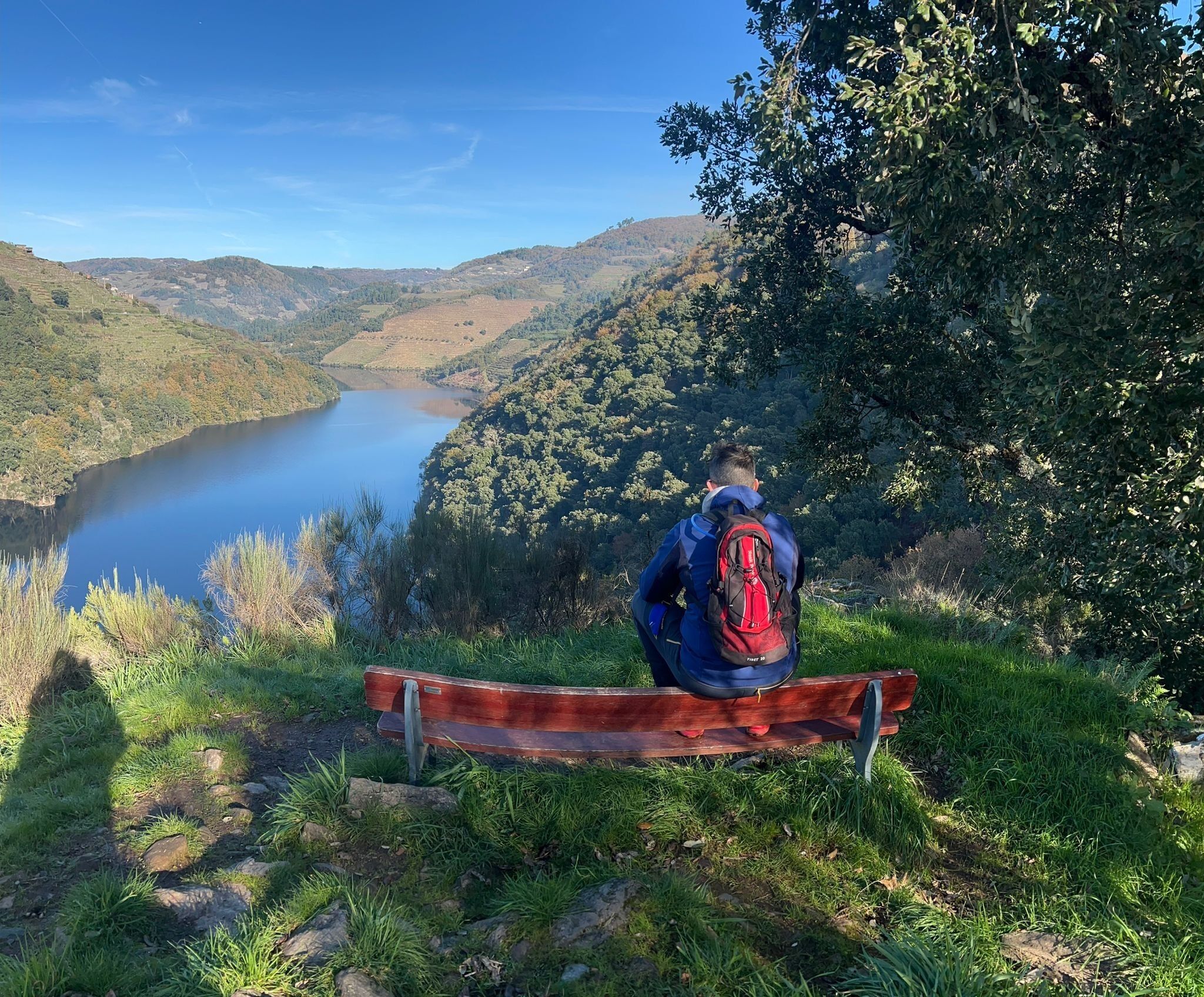 Eine Person sitzt auf einer Bank und blickt auf einen See und Hügel.