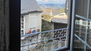 Una ventana con vistas a un paisaje montañoso, un balcón con barandilla metálica y un edificio con fachada de piedra.
