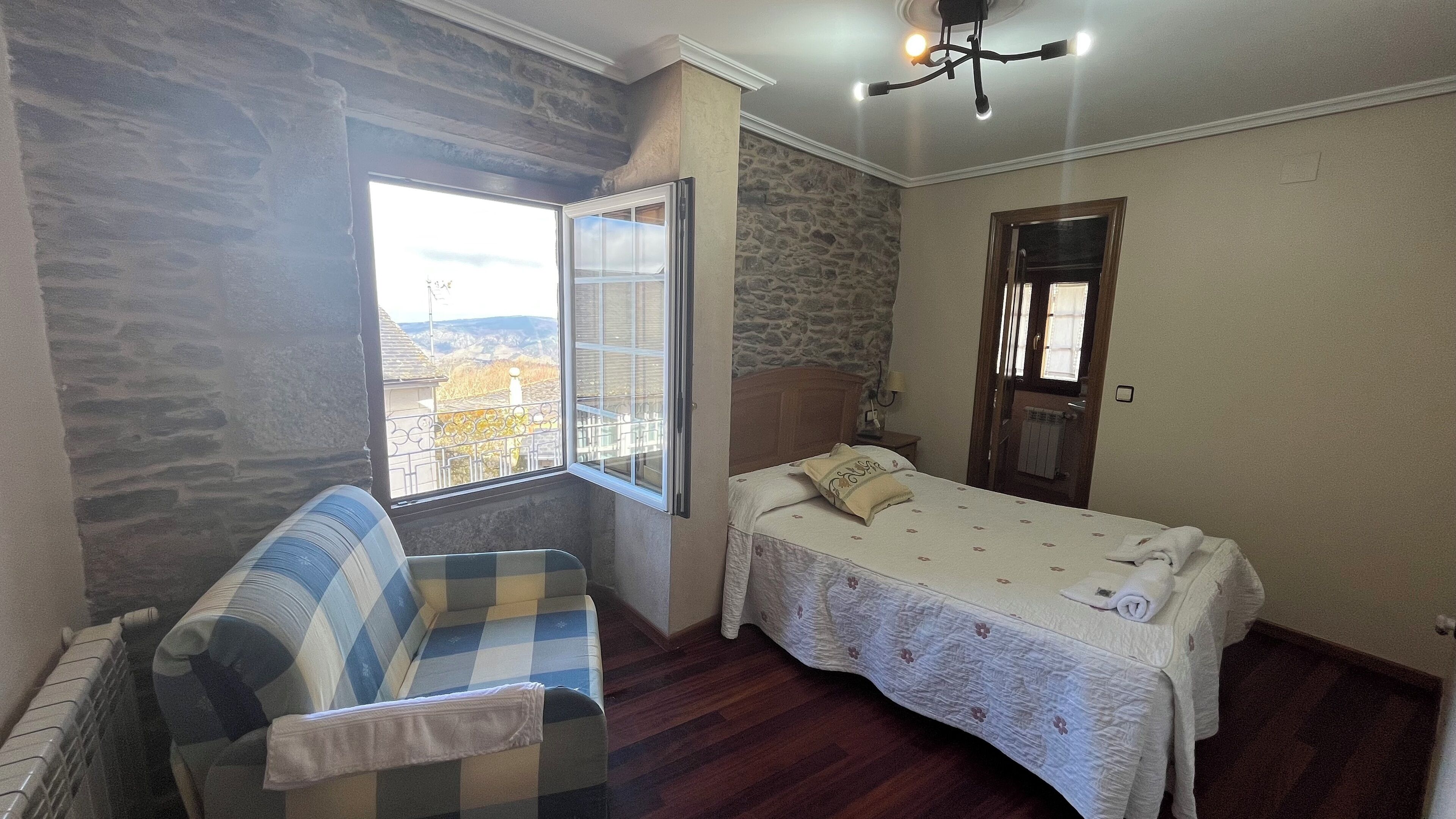 A bedroom with a bed, a window with a view, a sofa, and a stone wall.