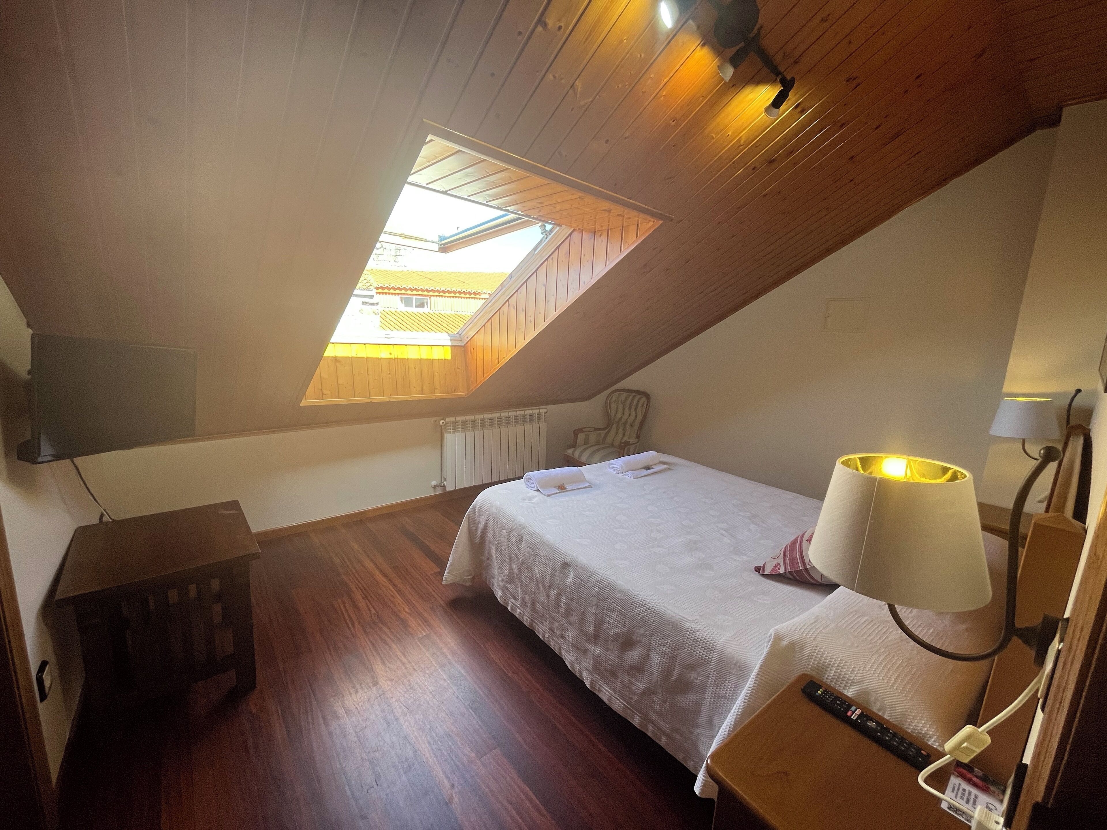 Un dormitorio con techo inclinado, tragaluz, cama con cubrecama blanco, lámpara de noche, mesita de noche de madera y una silla.