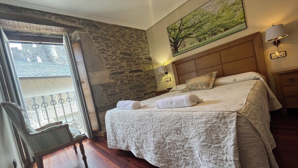 Habitación de hotel con pared de piedra, cama con cubrecama blanco, cabecera de madera, mesita de noche con lámpara y ventana con vistas a un edificio.
