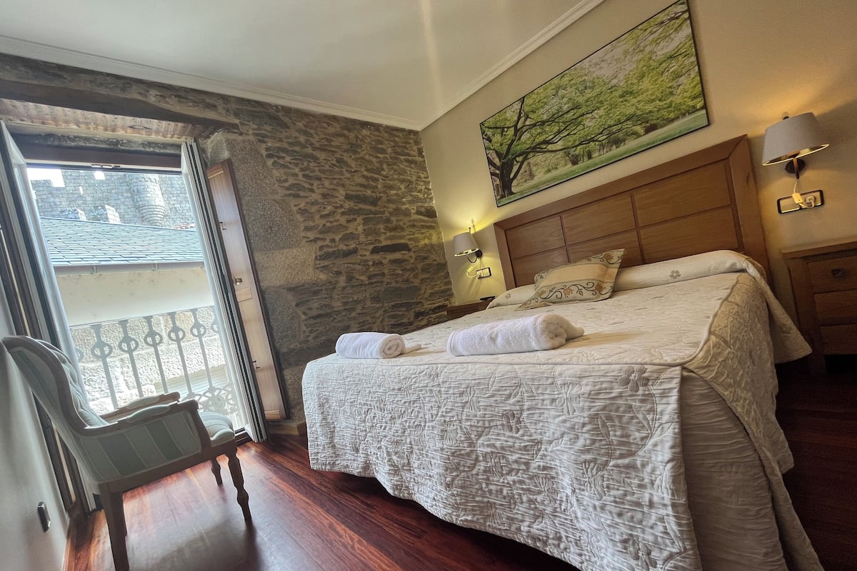 Quarto de hotel com parede de pedra, cama com colcha branca, cabeceira de madeira, mesa de cabeceira com abajur e janela com vista para um edifício.