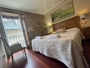 A hotel room with a stone wall, a bed with a white bedspread, a wooden headboard, a nightstand with a lamp, and a window with a view of a building. - Casa Alberto (Castro Caldelas)