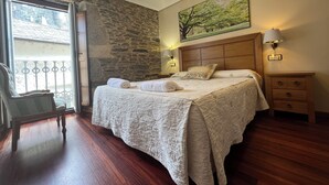 Un dormitorio con pared de piedra, cama con cabecera de madera, mesita de noche, una silla y una ventana con vistas a un edificio.
