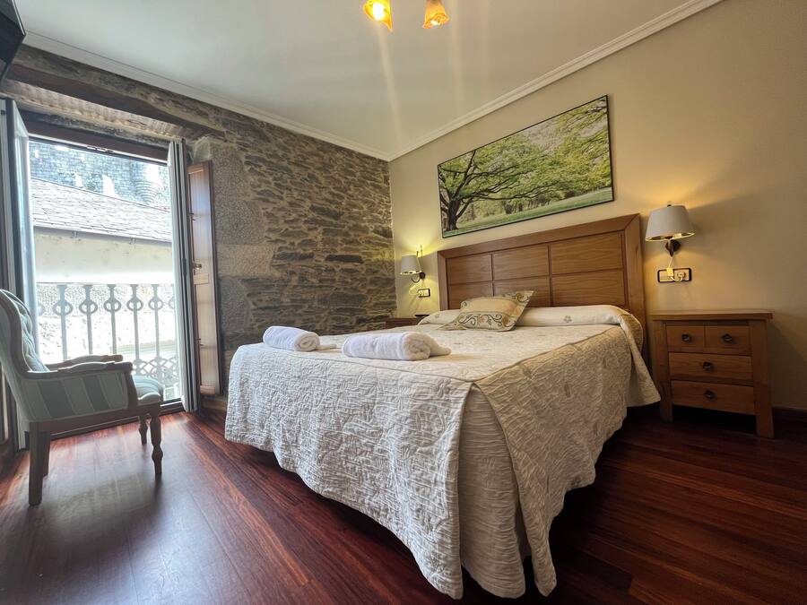 Un dormitorio con pared de piedra, cama con cabecera de madera, mesita de noche, una silla y una ventana con vistas a un edificio.